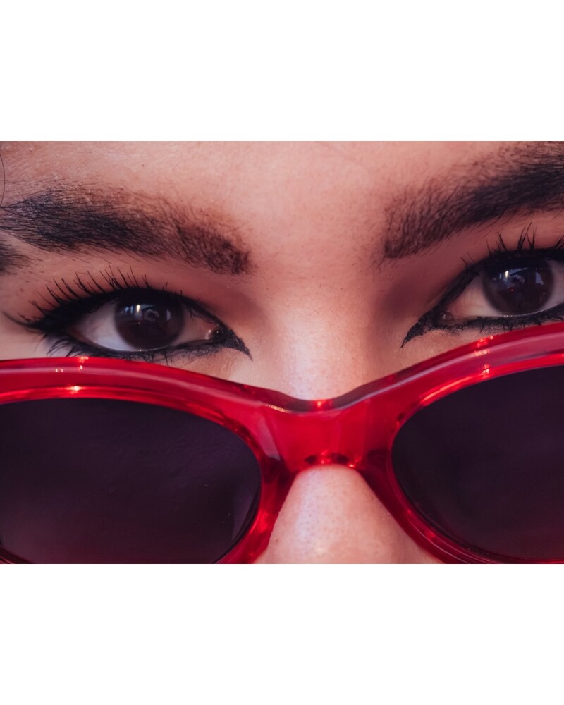 Close-up of a person's eyes peering over red cat-eye sunglasses with dark lenses and sharp winged eyeliner.