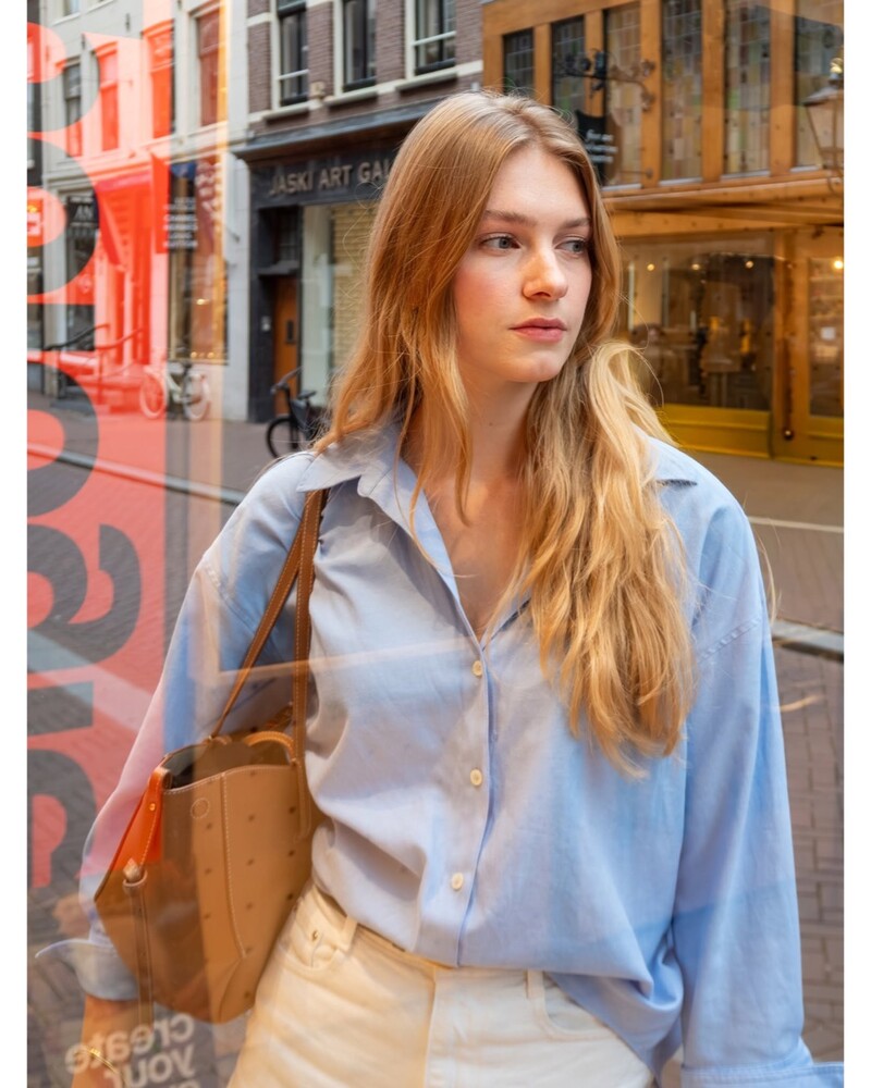 Woman with long blonde hair in a light blue shirt and tan tote bag, standing by a shop window on a city street.