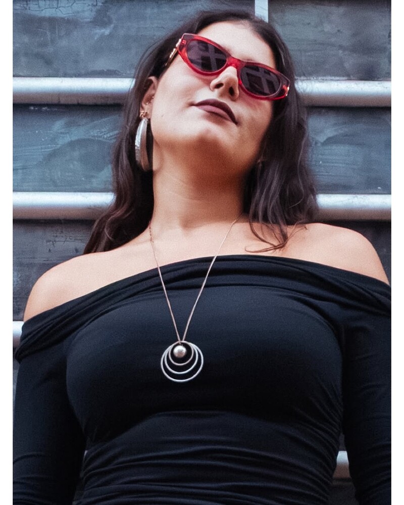Woman in a black off-shoulder top wearing red sunglasses, large silver hoop earring, and a layered circular pendant necklace, looking up against a gray corrugated wall.