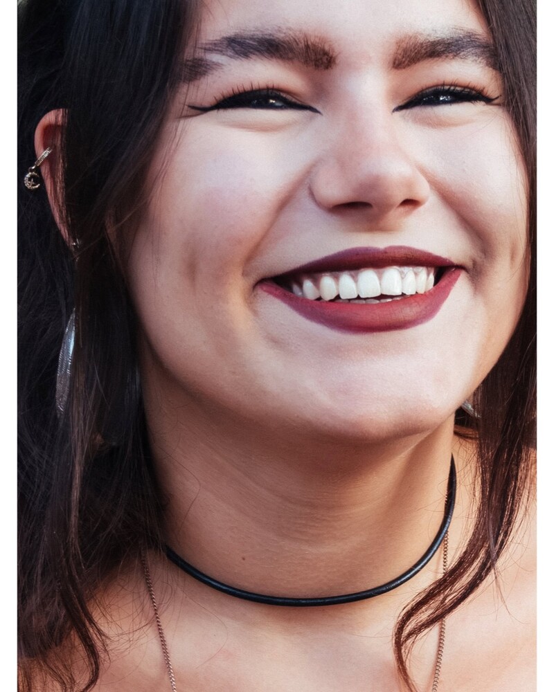 Close-up of a smiling woman with dark red lipstick, a black choker, and ear piercings against a blurred background