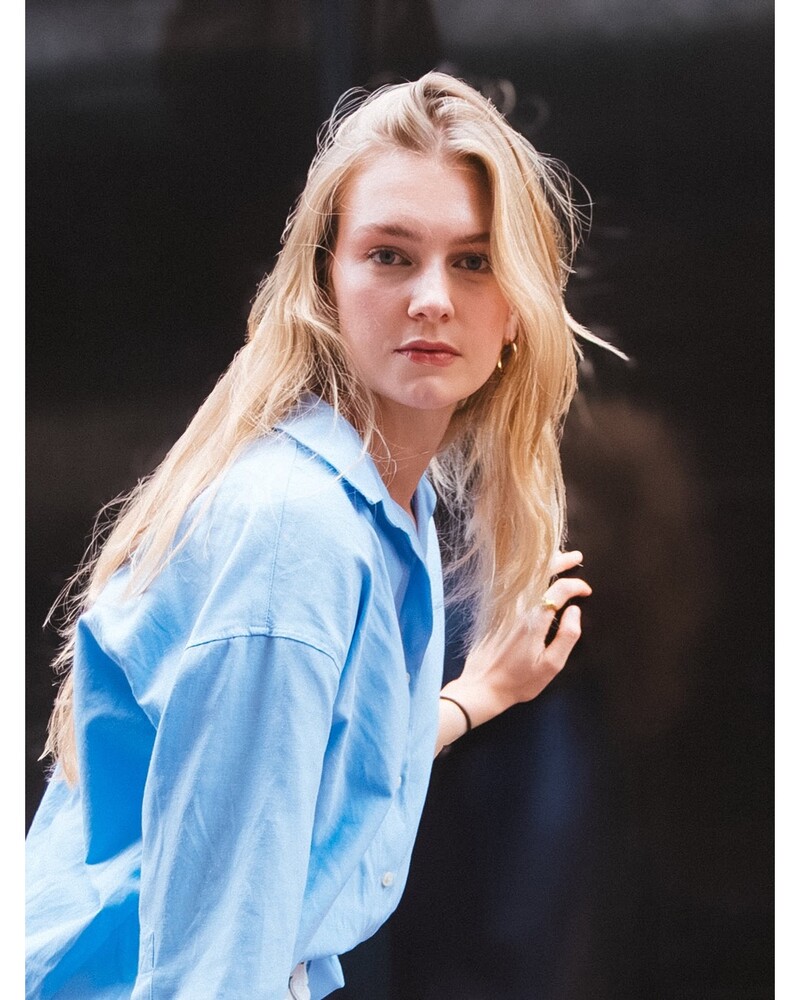 Woman with long wavy blonde hair in a light blue button-up shirt, glancing over her shoulder with one hand raised near her hair against a dark background.