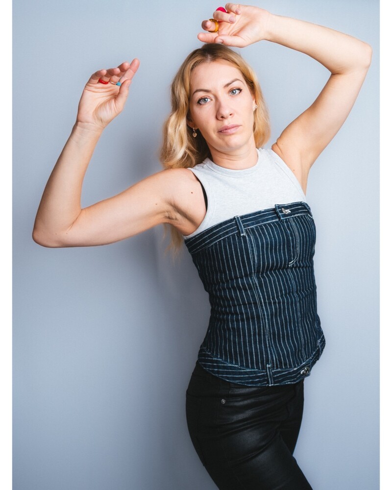 Blonde woman in a pinstripe corset top and black pants poses with both arms raised against a plain gray backdrop