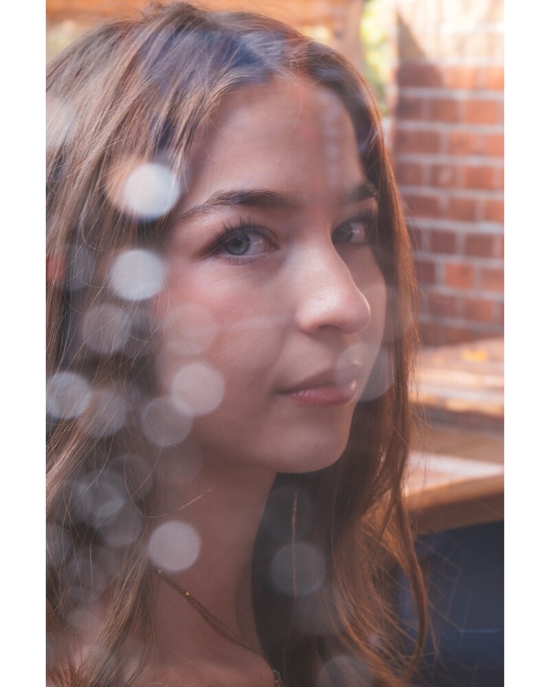 Young woman with long brown hair turning her face toward the camera, framed by out-of-focus bokeh orbs and a warm brick wall.