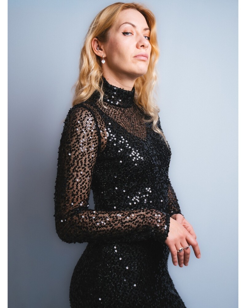 Woman with wavy blonde hair in a black sequined dress with sheer mesh sleeves, posed against a pale gray backdrop.