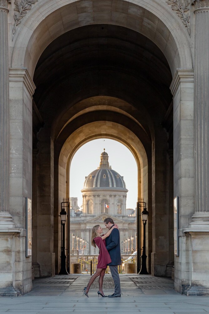 Couple embracing under a stone archway, with a domed building framed in the opening at golden hour in Paris.
