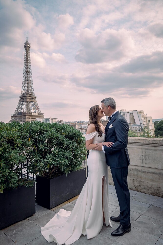 Bride and groom in a close embrace on a Paris rooftop terrace, with the Eiffel Tower rising behind them under a soft pastel sky.