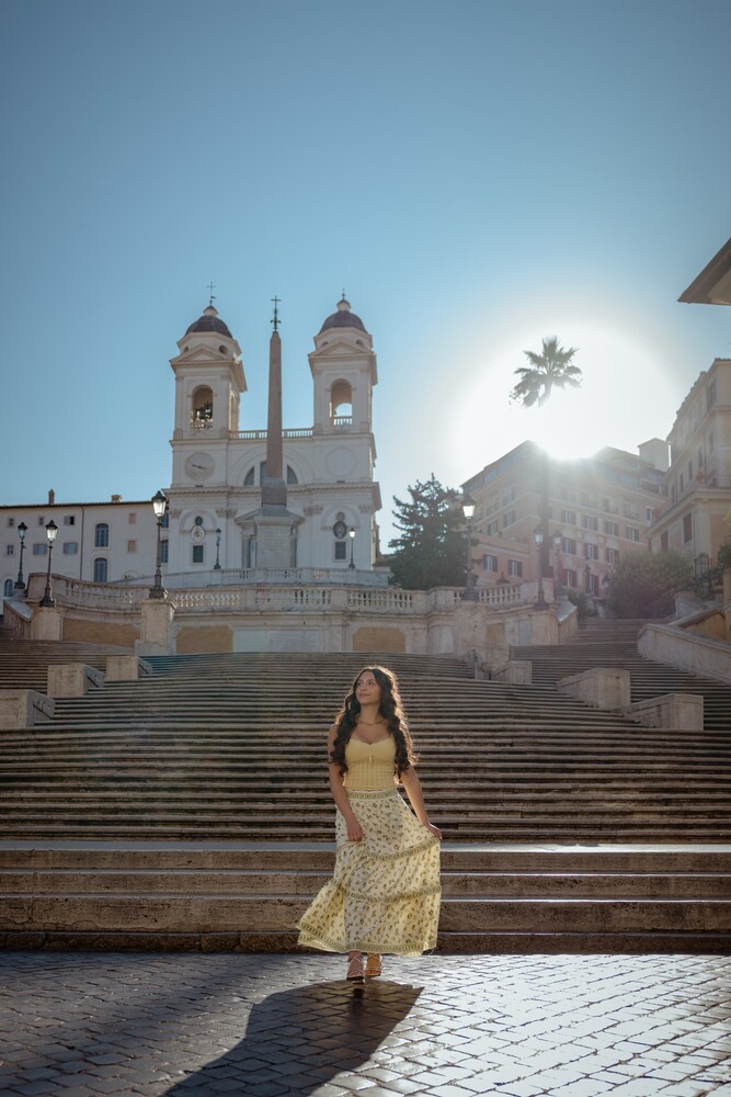 Woman in a yellow top and long patterned skirt standing at the base of Rome's Spanish Steps, with a backlit church and sun flare above.