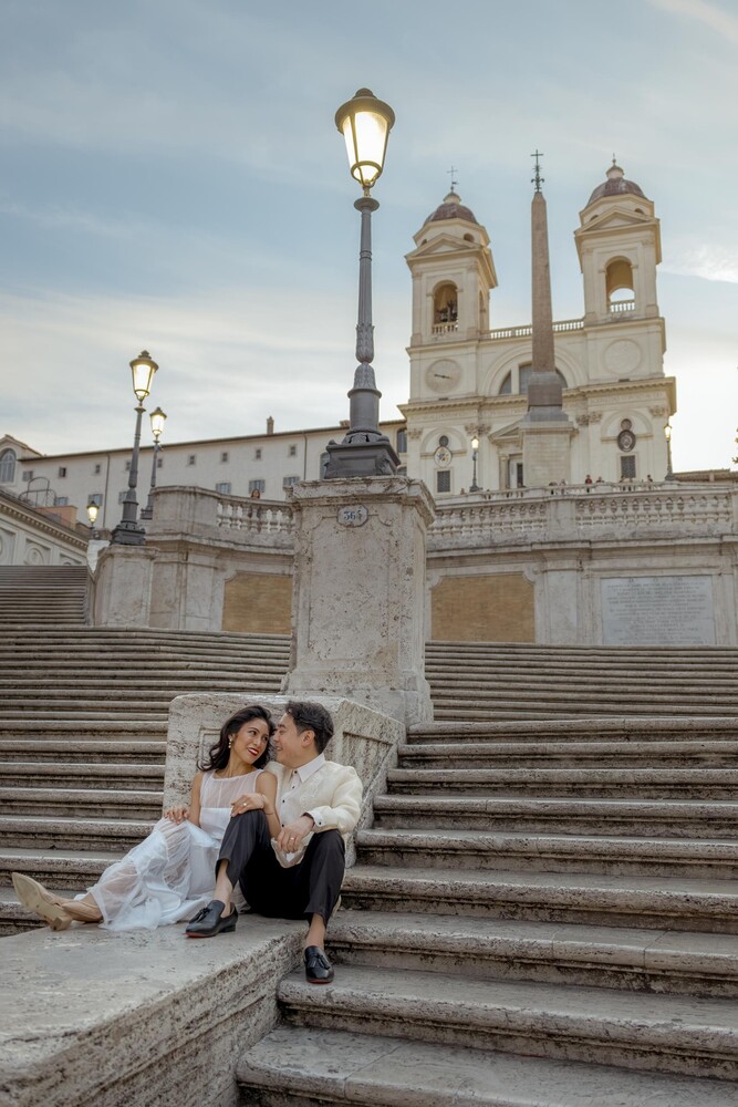 Couple embracing on the Spanish Steps in Rome, with the Trinità dei Monti church and an ornate street lamp behind them