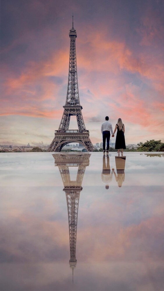 Couple holding hands on a reflective wet plaza, facing the Eiffel Tower at sunset under a pink and purple sky.