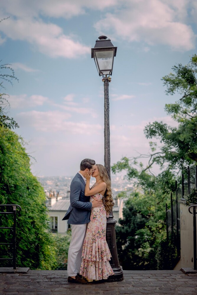 Couple embracing and kissing beside an old iron street lamp on a stone staircase, with a hazy city view and green trees behind them.