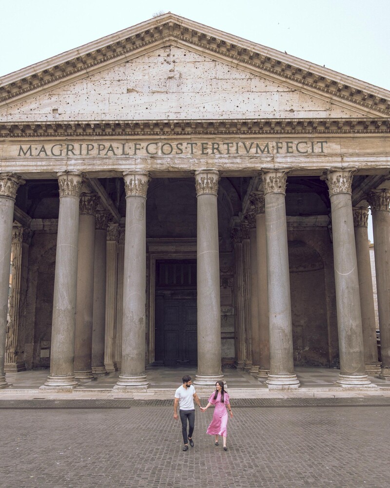 The Pantheon in Rome with a couple in pink and white walking hand in hand across the plaza toward its columned portico