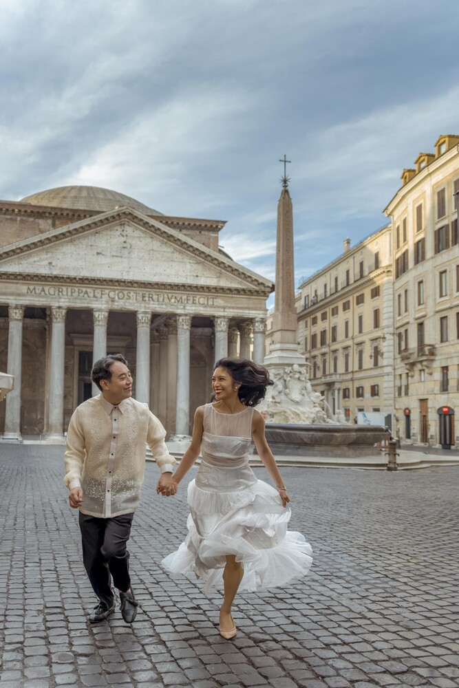Bride and groom run hand in hand across a cobblestone square toward the Pantheon in Rome.