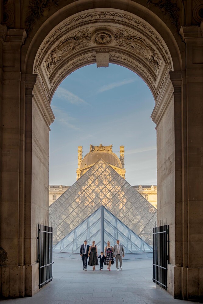 View through an ornate stone archway of the glass pyramid at the Louvre in Paris, with small groups of visitors walking across the courtyard.