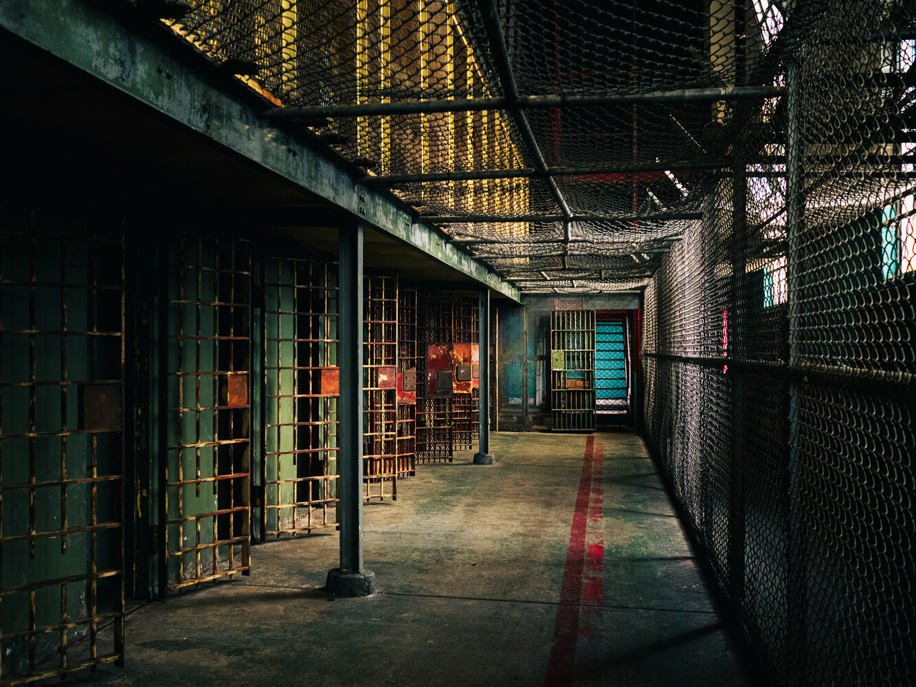 Prison corridor with barred cell doors on the left, chain-link fence on the right, and a red line leading to a teal door.