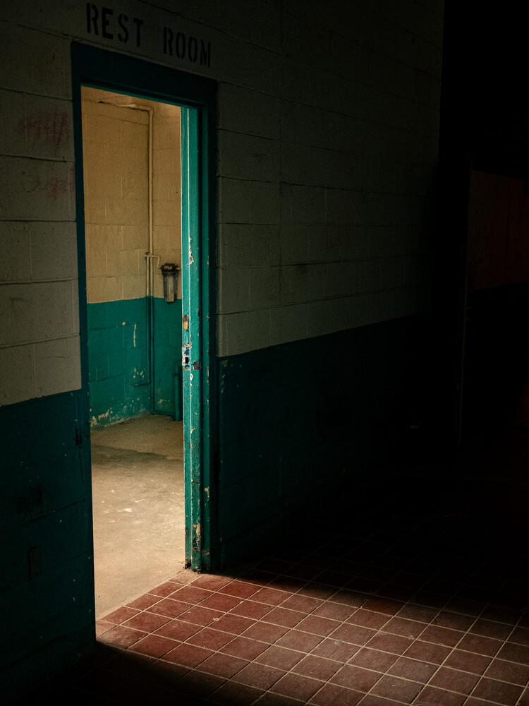 Open teal doorway leading into a dimly lit restroom, with a red tile floor and a REST ROOM sign above on a painted block wall.