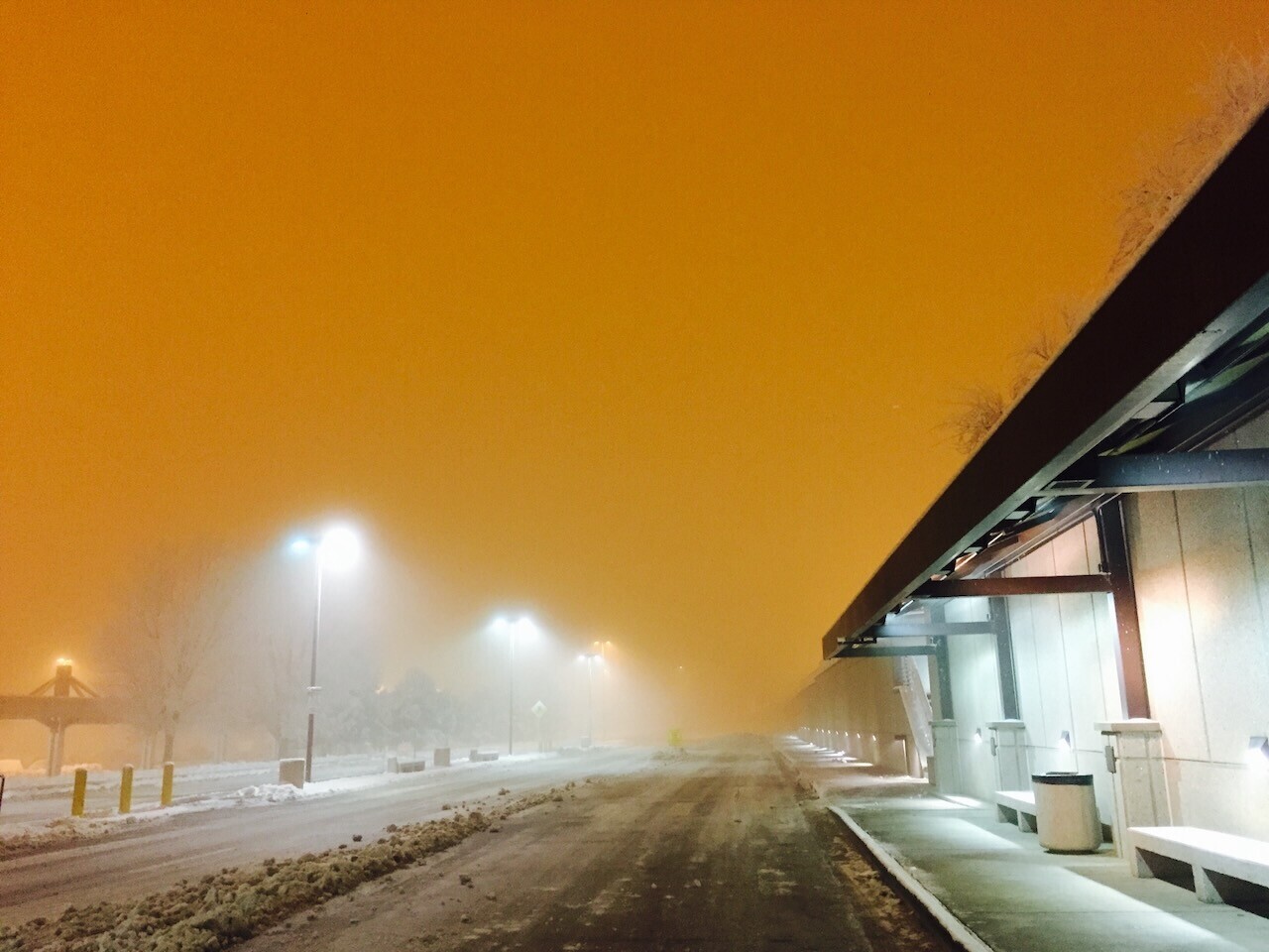 Snowy road at night under an orange sky, street lamps glowing through fog and falling snow beside a covered walkway.