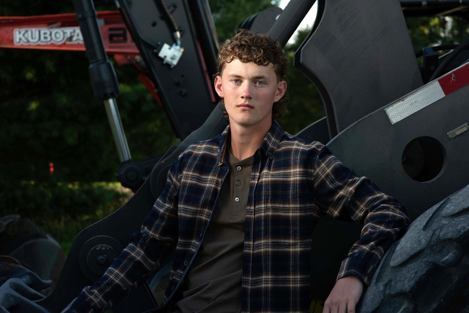 Young man in plaid shirt leaning against farming equipment outdoors