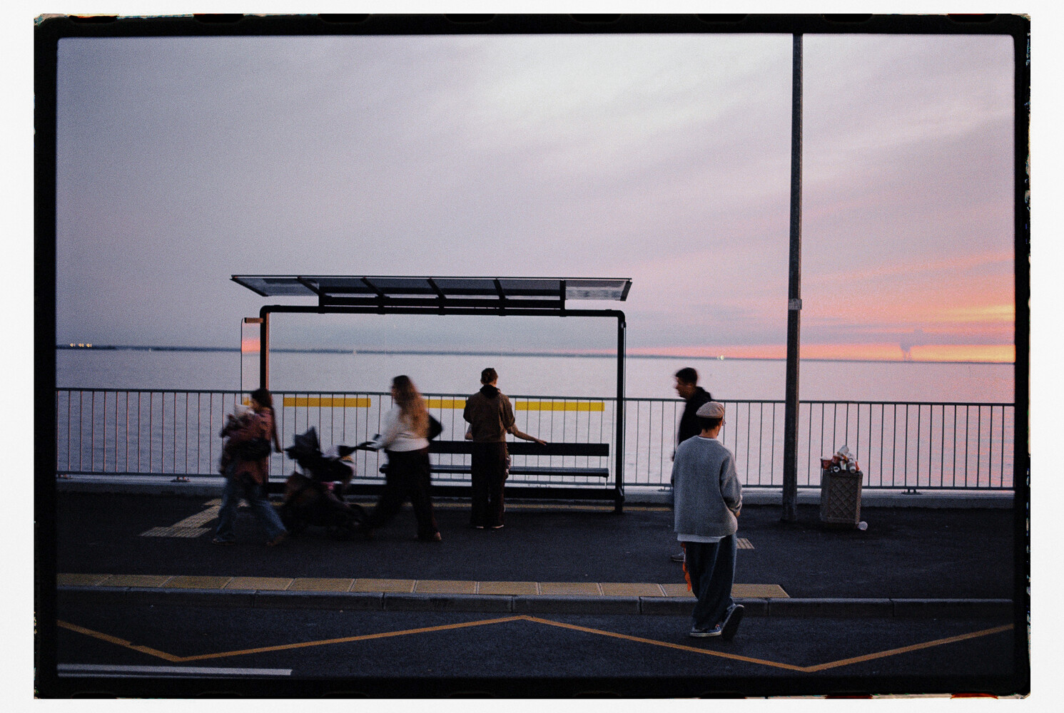 People gathered at a waterfront bus shelter at sunset with calm water and pink sky in background