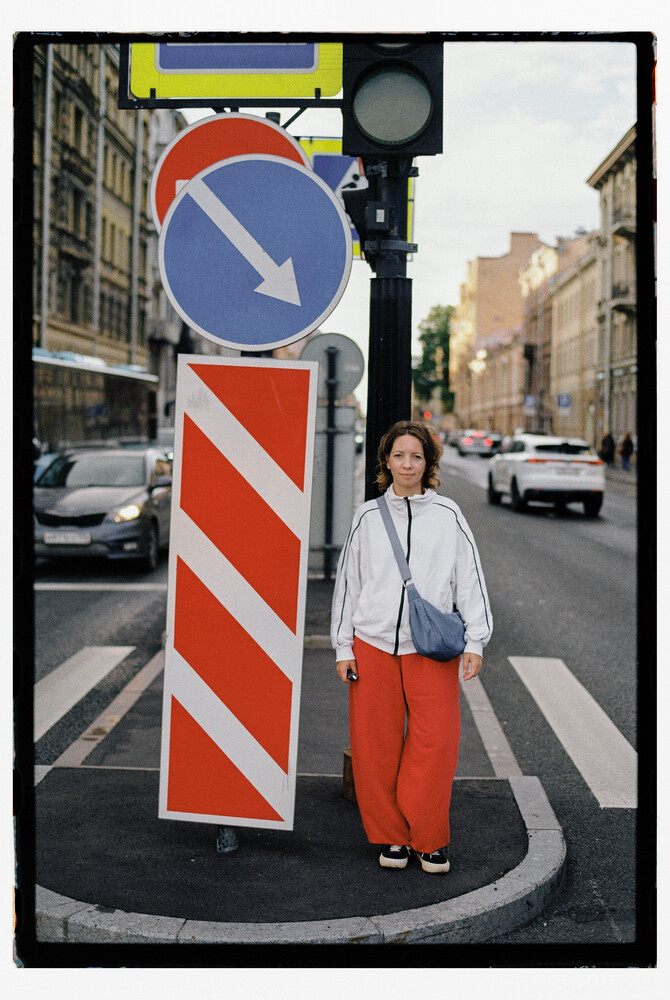 Woman in white shirt and red pants standing beside blue directional traffic sign with red diagonal stripes on urban street
