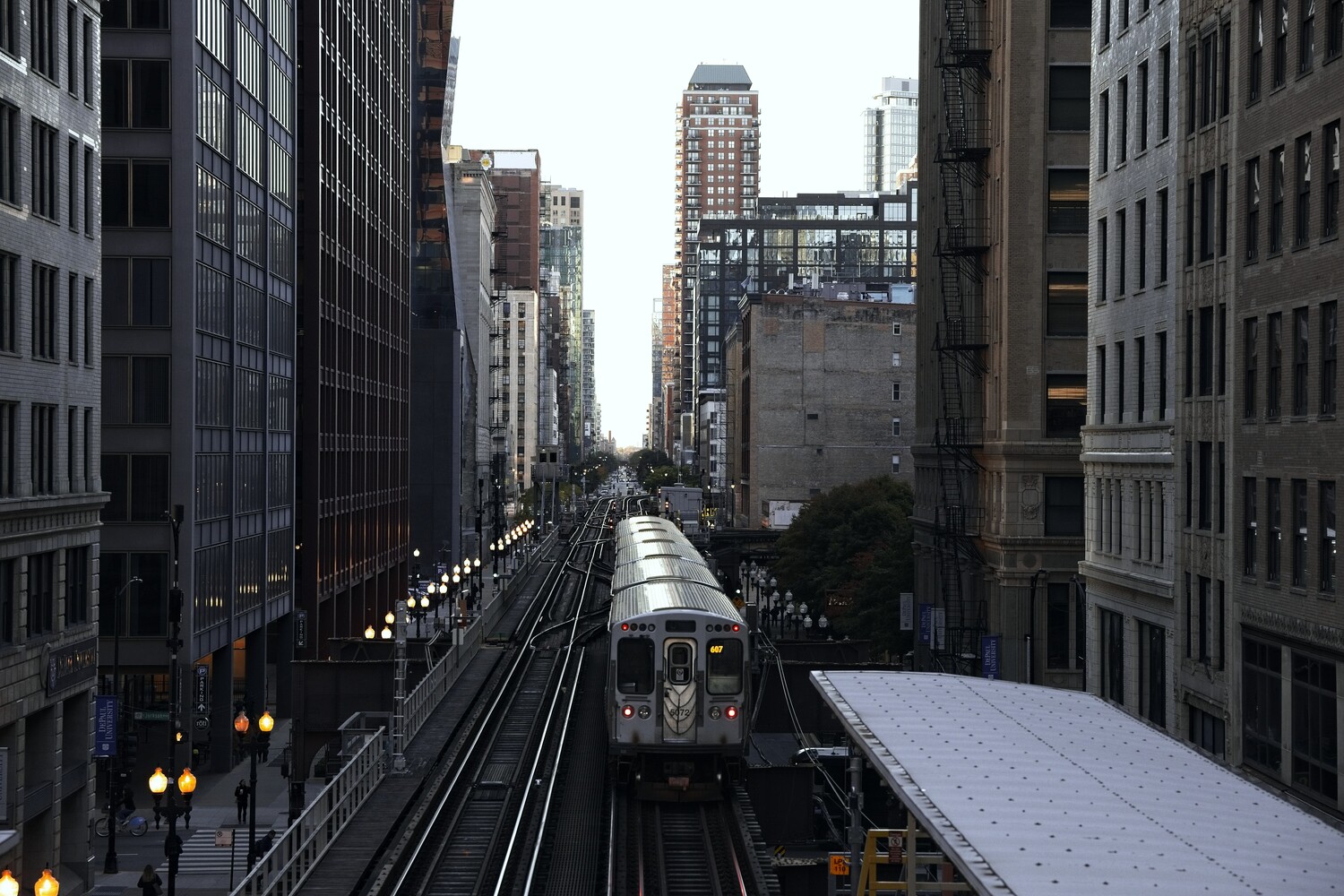 Silver elevated commuter train approaching down a steel rail corridor, framed by tall city buildings on both sides