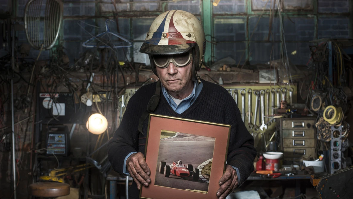 Craftsman in a workshop holding a framed photograph of a race car, surrounded by hanging tools and equipment.