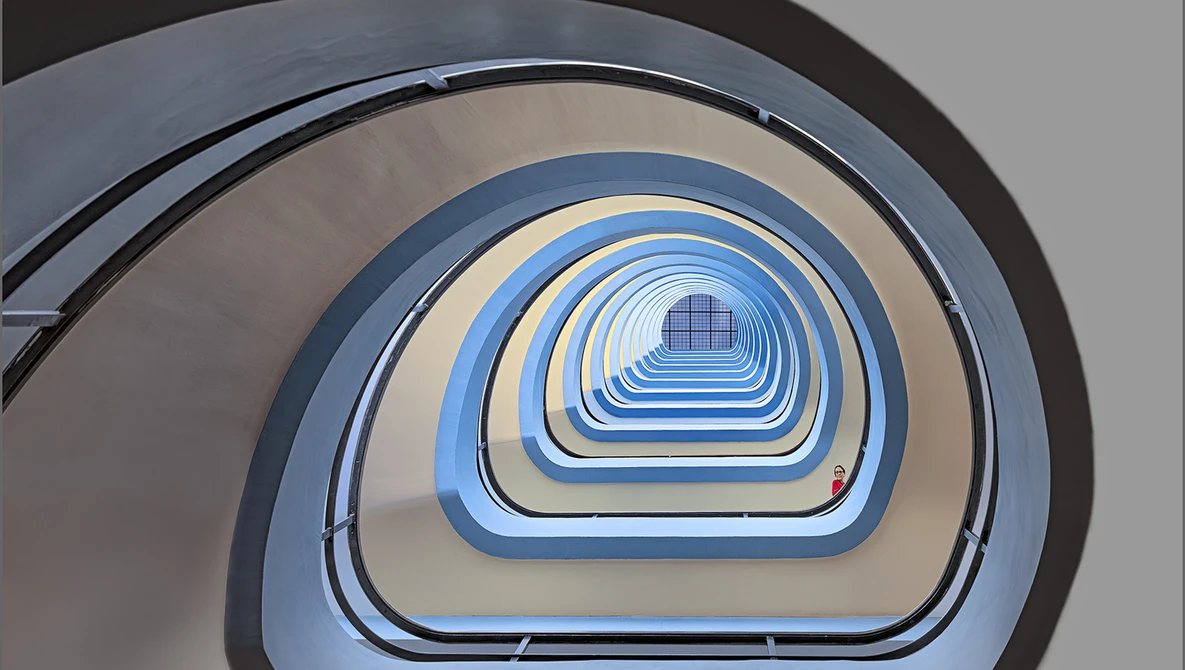 Overhead view of a spiral staircase descending through concentric rounded rectangular frames in blue, beige, and gray tones.