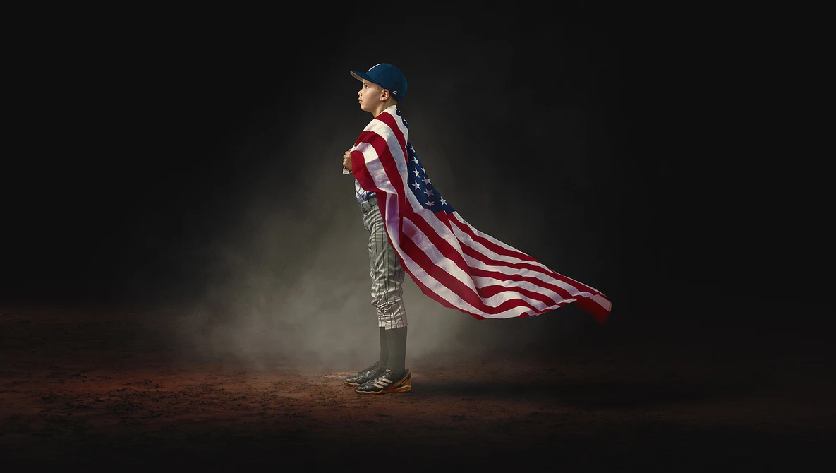 Cowboy in hat and vest holding an American flag that billows behind him against a dark background.