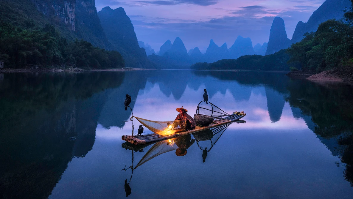 Fisherman in a small boat with a glowing lantern on a mirror-still river at twilight, surrounded by dramatic karst mountains.