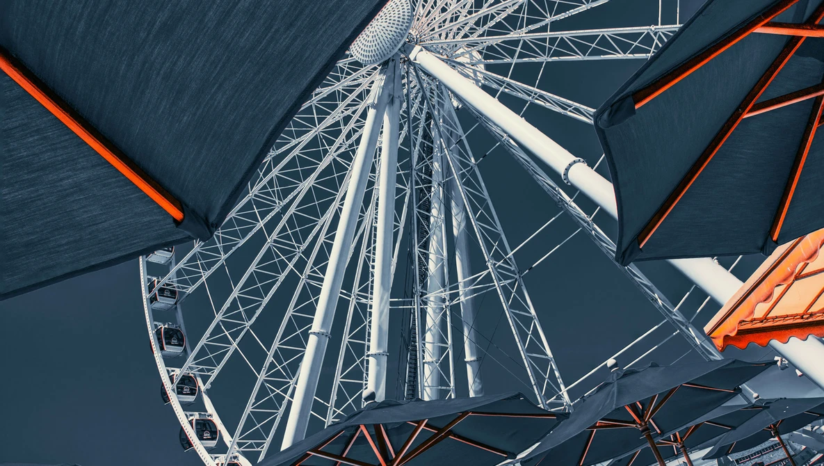 Upward view of a Ferris wheel's intricate white metal framework against a dark blue sky, with orange structural supports visible on either side.