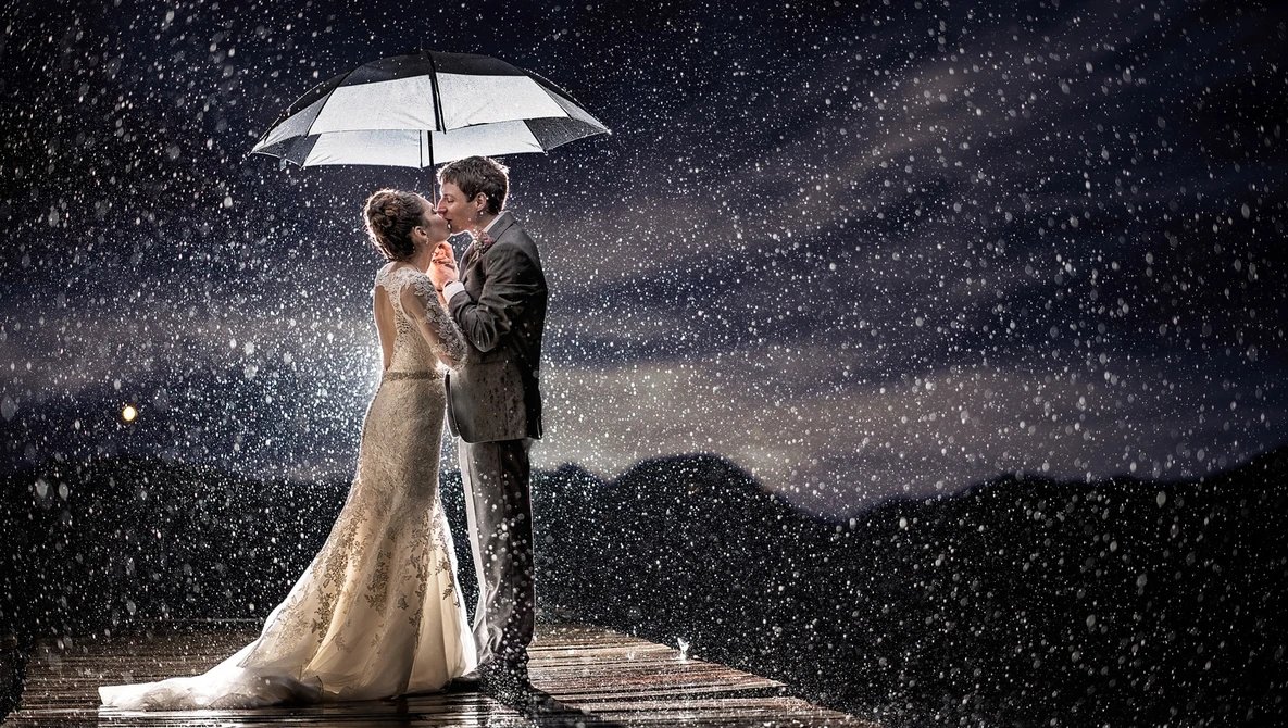 Bride and groom kissing under a white umbrella in heavy rain at night.