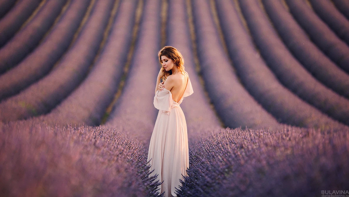 Woman in white backless gown standing in lavender field rows at golden hour.
