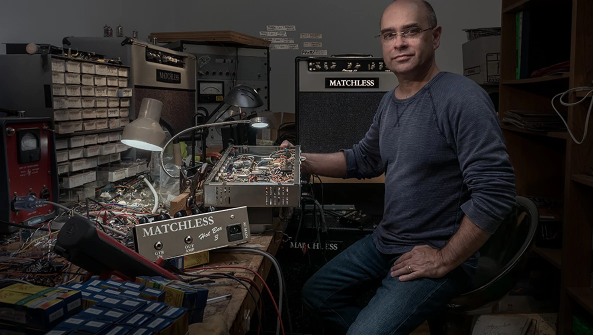 Man in home electronics workshop holding circuit board with soldering equipment visible.