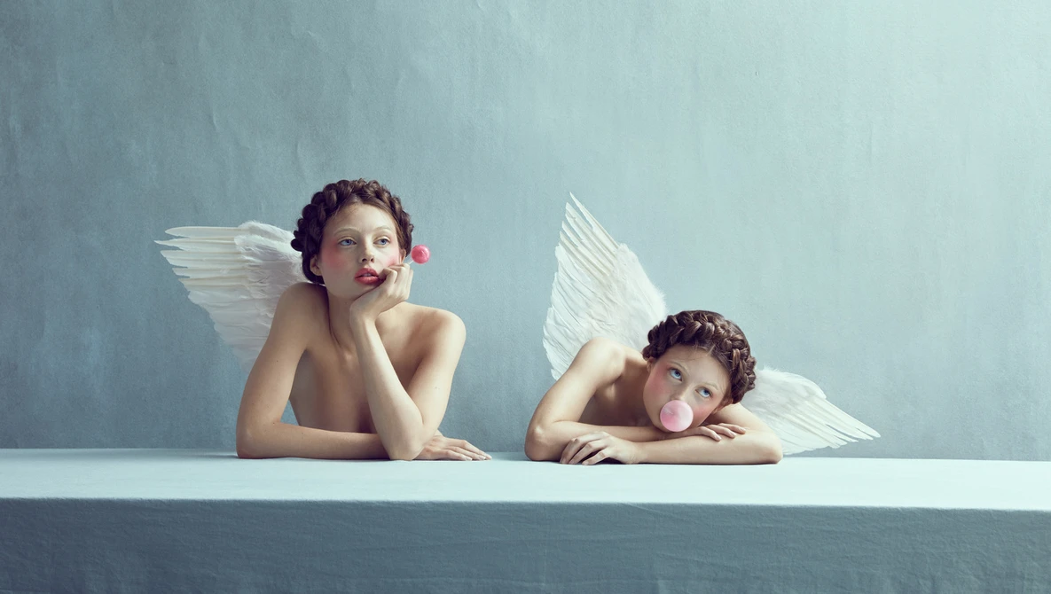 Two figures with white wings posed against a muted blue-gray backdrop, one seated upright and one reclining on a white surface.