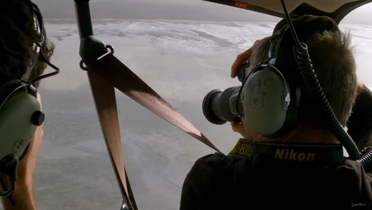 Aerial photographer with Nikon camera shooting from a helicopter, capturing landscape below through open door.