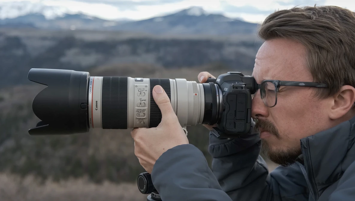Photographer with glasses holding a telephoto lens camera, shooting toward distant snow-capped mountains.
