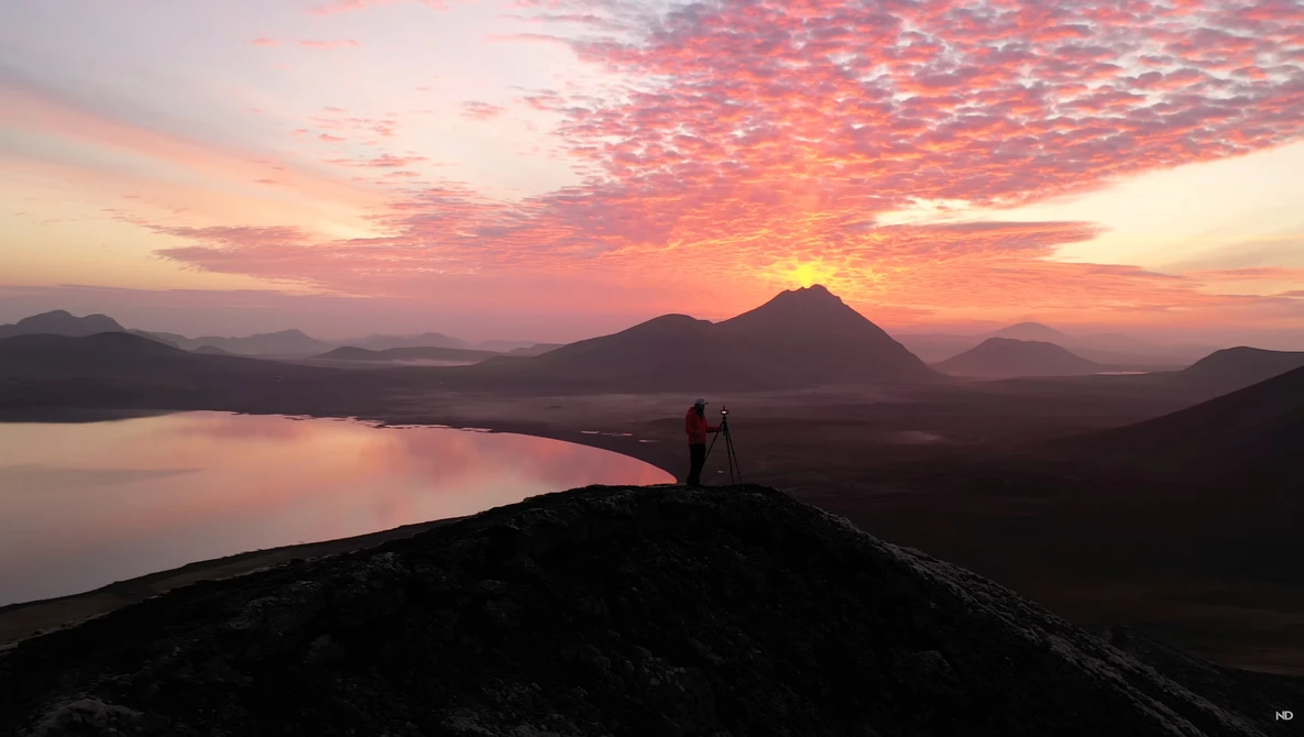 Silhouetted figure standing on a dark ridge overlooking a lake at sunset with volcanic peaks in the distance.
