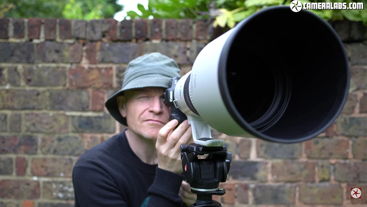 Photographer holding a large telephoto lens mounted on a camera with tripod head, wearing a cap against a brick wall background.