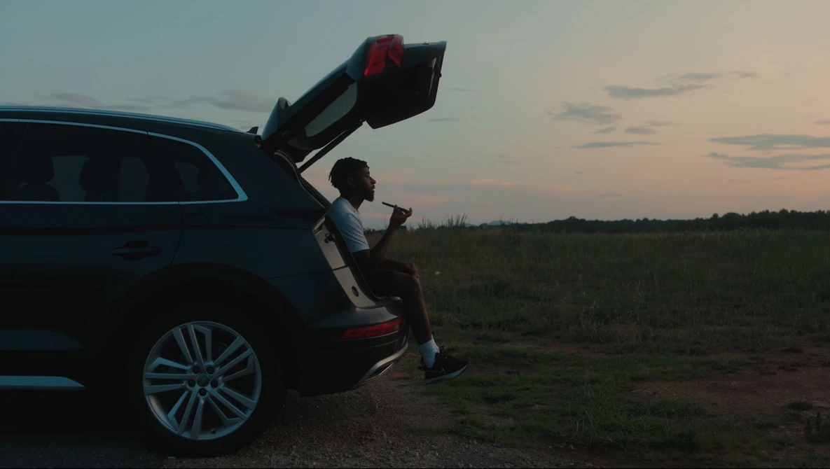 Person sitting in open car trunk at dusk in an open field, gazing toward the horizon.