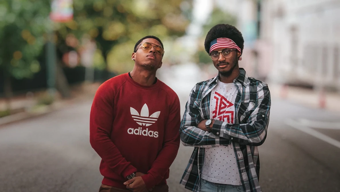 Two men standing on a street wearing casual sportswear and sunglasses.