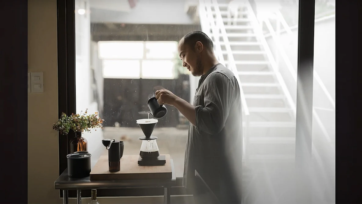Man in casual shirt preparing pour-over coffee near a bright window with natural morning light.