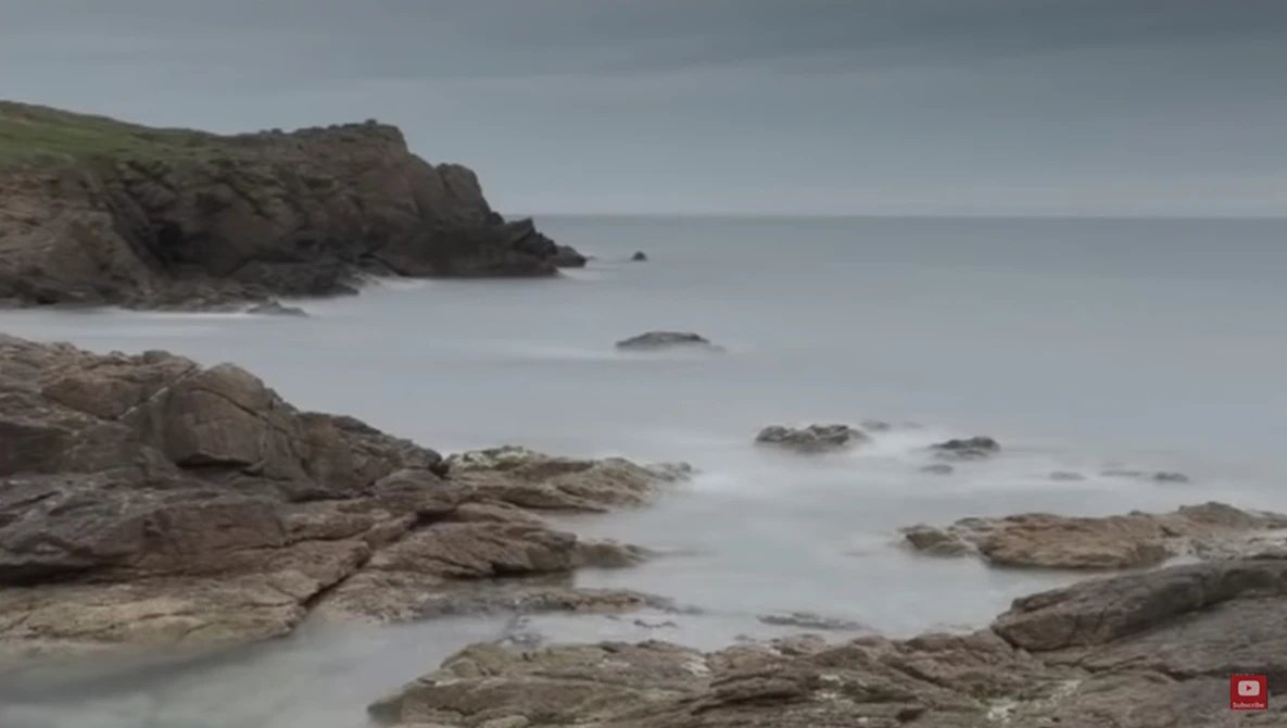 Rocky coastal landscape with smooth water and misty headland under overcast sky.