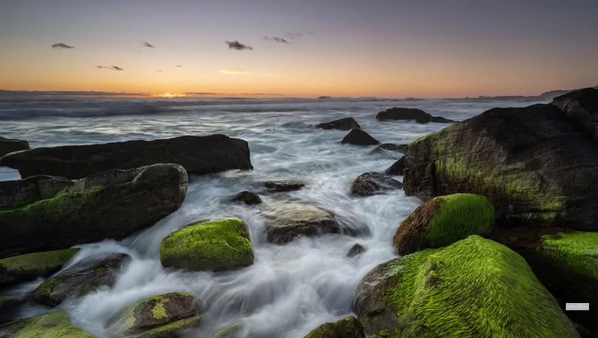 Coastal landscape at sunset with moss-covered rocks and flowing water in foreground.