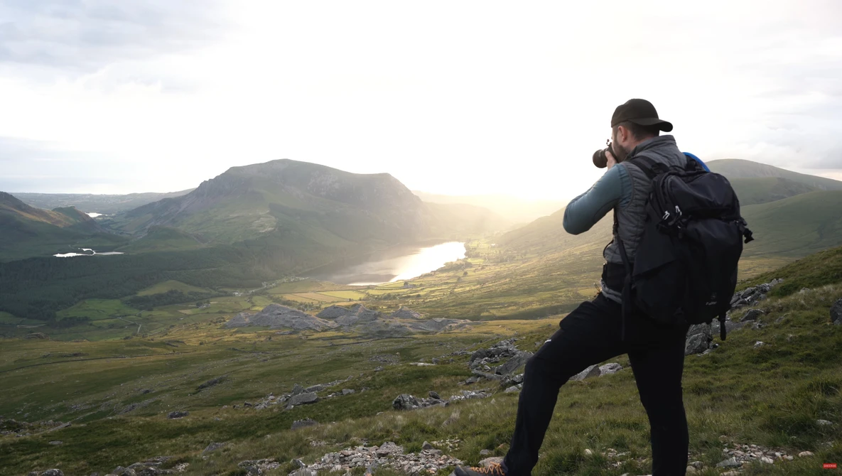 Photographer with camera and backpack capturing landscape from hilltop at golden hour.