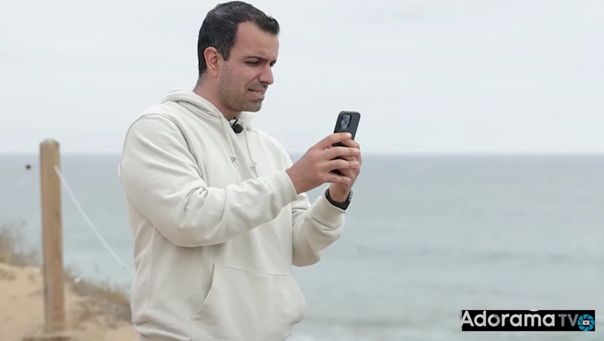 Man in beige hoodie holding smartphone while standing on beach with ocean and wooden post in background.
