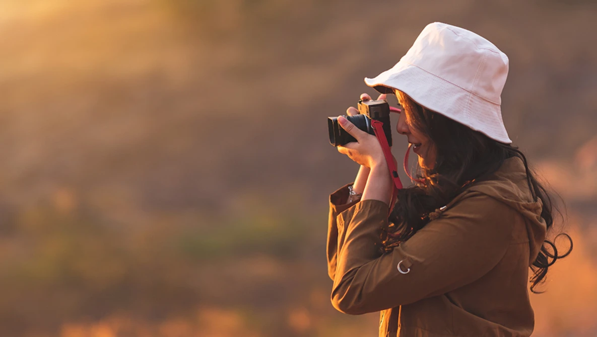 Photographer wearing a white bucket hat and brown jacket holding a camera up to their face during golden hour.