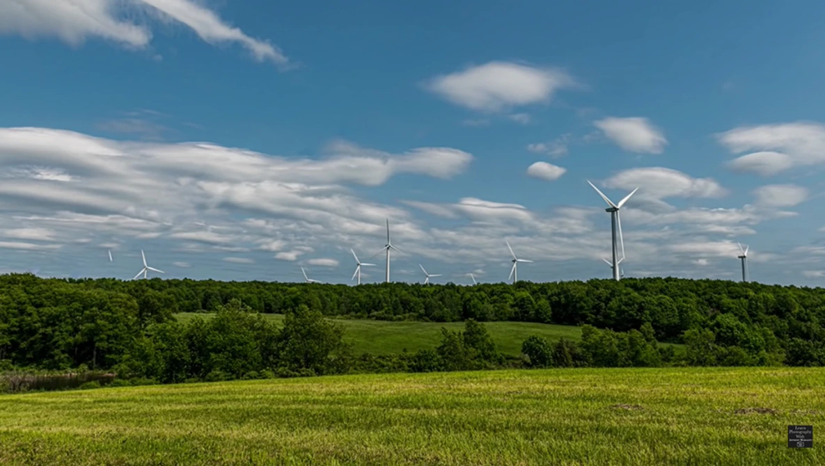Wind turbines dotting a forested landscape under a partly cloudy sky.