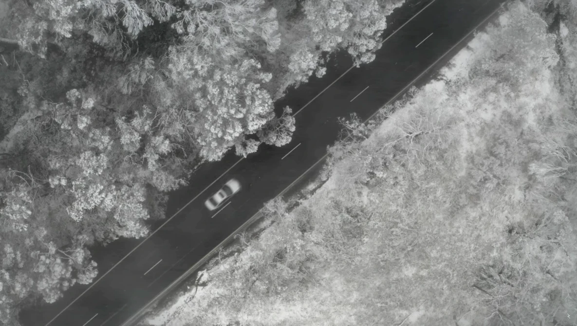 Aerial view of a vehicle driving on a dark asphalt road cutting through snowy forest terrain.