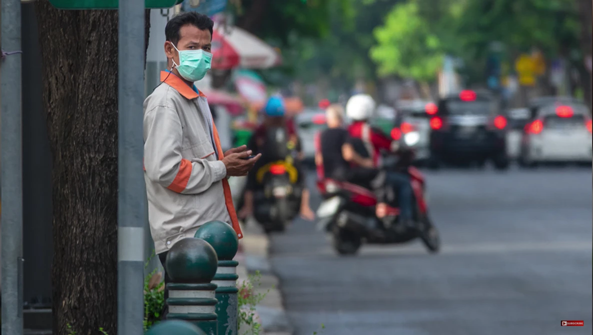 Man wearing surgical mask and orange safety vest standing against pole on urban street.