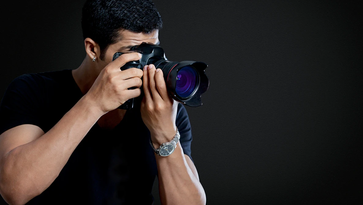 Male photographer holding a professional DSLR camera with purple lens element visible, shot against dark background.