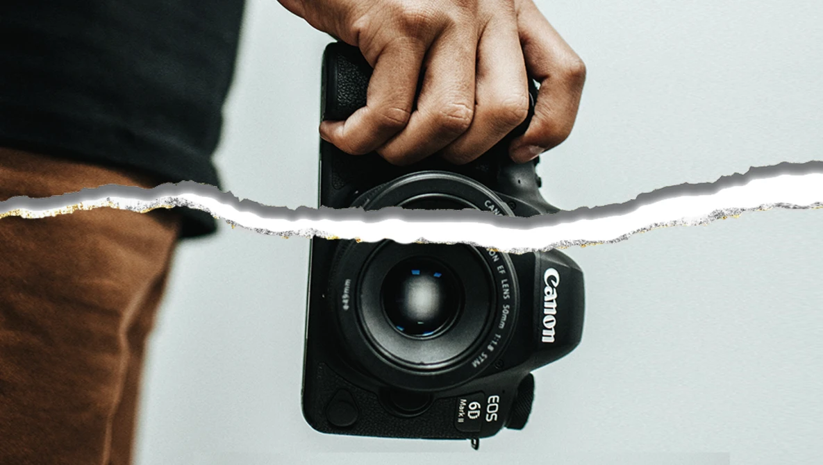 Photographer's hands holding a Canon EOS camera with a torn white paper overlay obscuring the upper portion.